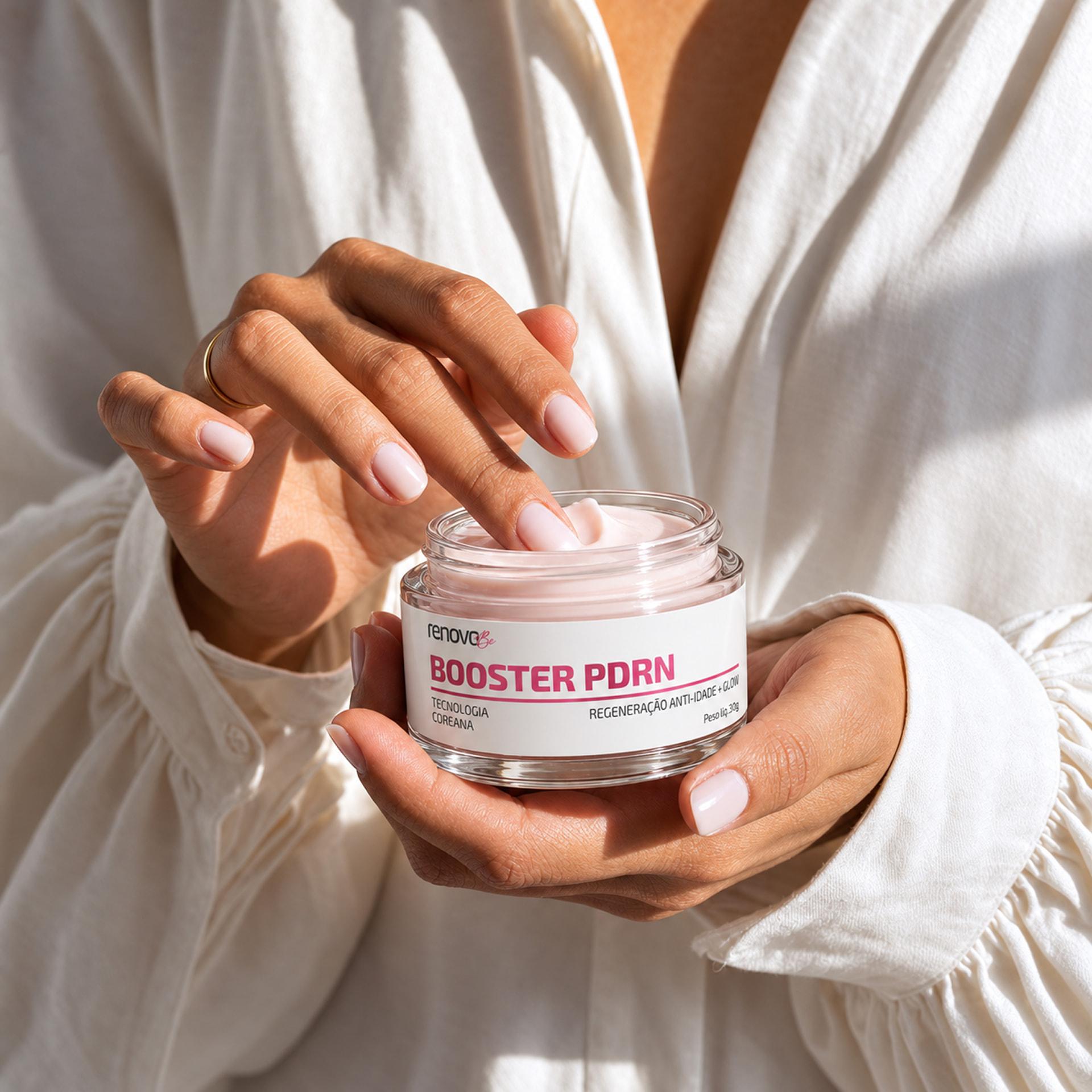 Mãos segurando o pote aberto do Booster PDRN da Renova Be, com o dedo no creme rosa